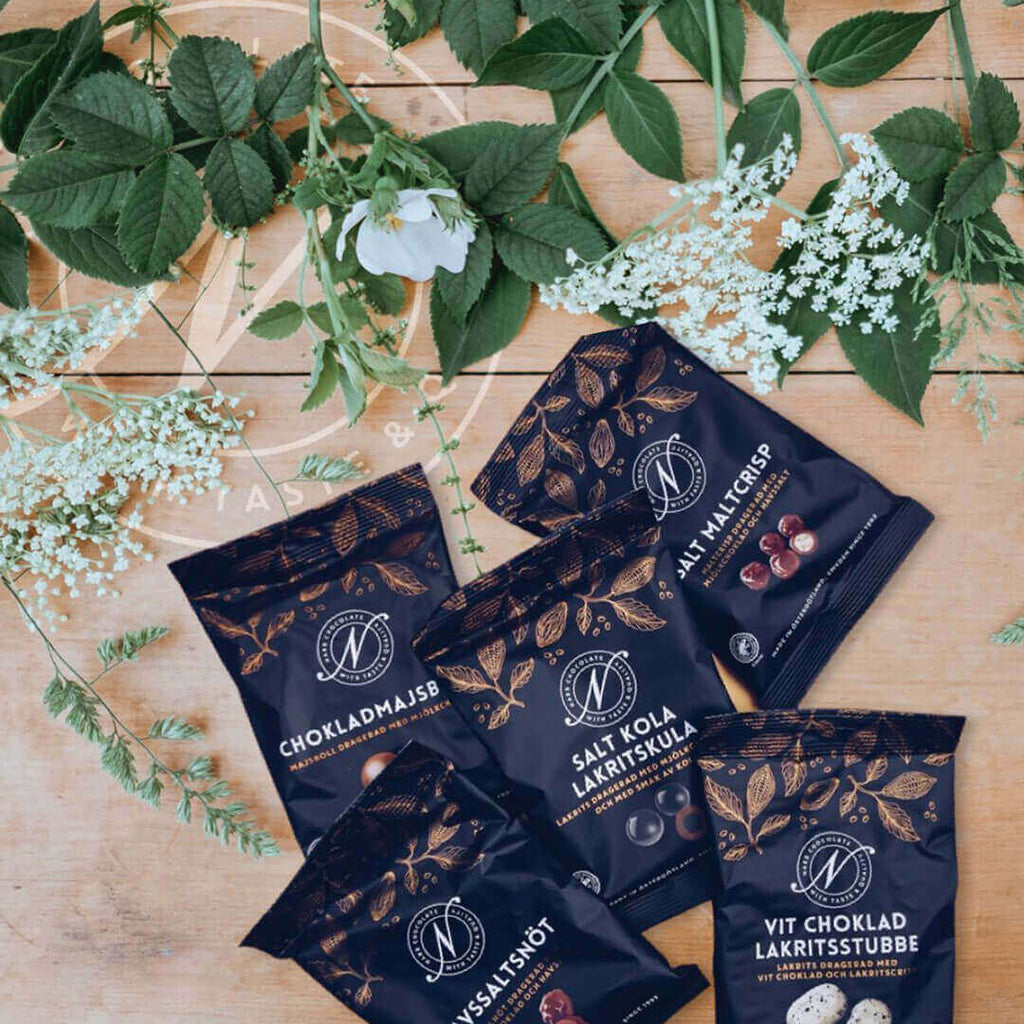 Fem påsar från Narr Chocolate på ett träbord med gröna blad och fläderblommor, bland dem Vit choklad lakritsstubbe, Salt kola lakritskula och Havssaltsnöt.
