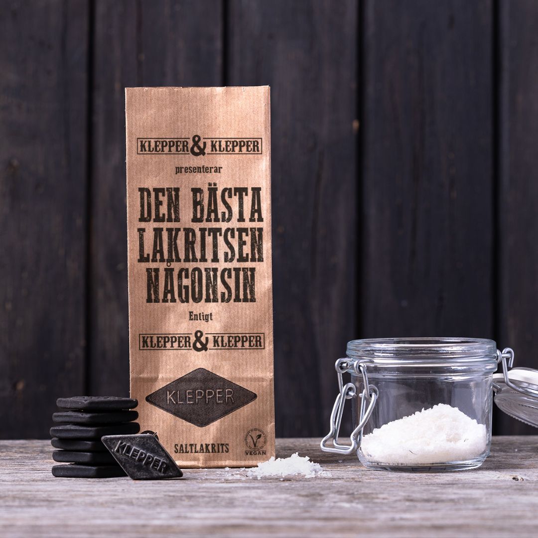 Påse saltlakrits från Klepper & Klepper på ett träbord, bredvid rombformade lakritsbitar och en glasburk med havssalt.